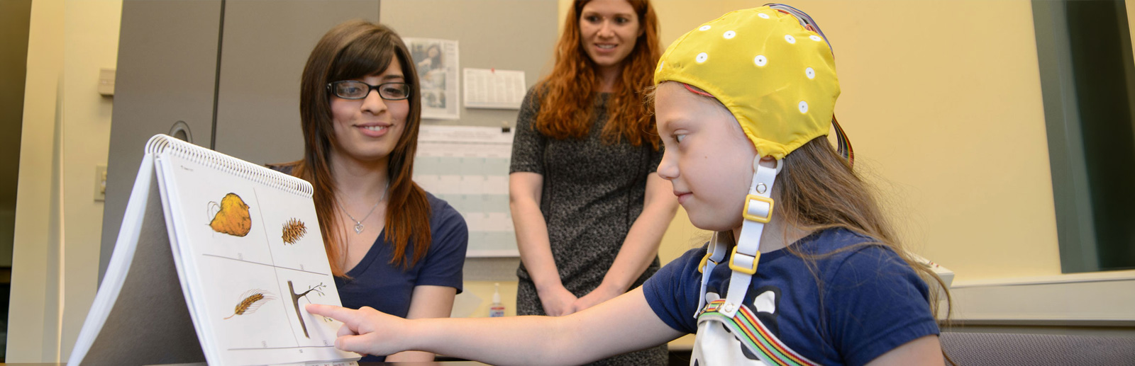 Child wearing a yellow brain cap pointing on one of four objects on a paper while two adults look on. 